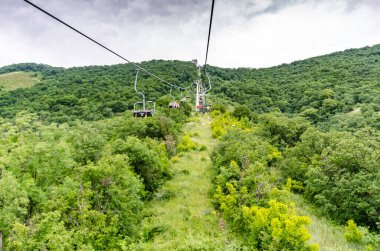 Yoğun orman tarafından çerçevelenmiş, yemyeşil tepeler üzerinde kablo araba süzülüyor. Bulutlu gökyüzü canlı manzaraya kontrastlık ekler. Bakış açısı huzurlu, doğaya yüksek bir bakış açısı sunar