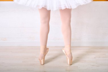 Beyaz etekli genç bayan bacaklar nokta tekniği uyguluyor. Aydınlık ahşap zemini ve yumuşak ışıklandırmasıyla bale stüdyosunda duruyor. Hassas duruş zarafet ve disiplini vurgular.