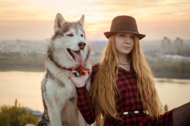 Kırmızı tişörtlü ve şapkalı genç kadın tepede Husky 'nin yanında duruyor. Günbatımı şehrin siluetini ılık tonlarda yıkar ve sakin bir atmosfer yaratır. İkisi de sakin ve hoşnut görünüyor.