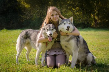 Uzun kızıl saçlı genç bir kadın güneşli parkta iki köpeğe sarılıyor. Yeşil çimenler çevrelemiş, ağaçlar gölgeli bir zemin oluşturmuş. Sıcak, neşeli sahne yoldaşlığı ve doğayı yakalar.