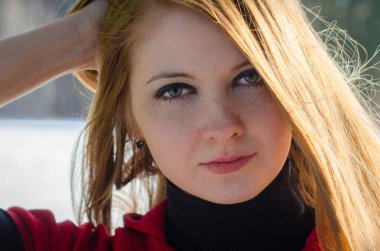 Kızıl saçlı ve çilli genç bir kadın kendinden emin bir şekilde bakıyor. Parlak güneş ışığı, yumuşak bulanık arka plana karşı onun özelliklerini vurgular. Siyah ve kırmızı elbiseler giyiyor.