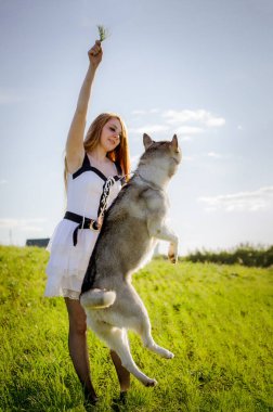 Uzun kızıl saçlı genç bir kadın çimenli bir tarlada duruyor, dalları şakacı bir şekilde kabuğunun üzerinde tutuyor. Parlak güneş ışığı yumuşak gölgeler oluşturur. Arka plandaki uzak yapı derinlik ekler.