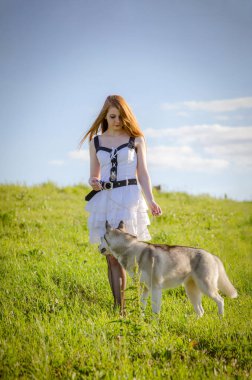 Uzun kızıl saçlı ve beyaz elbiseli genç bir kadın yeşil çayırda Husky 'nin yanında duruyor. Parlak öğleden sonra güneşi kırsal araziyi aydınlatır. Gökyüzünde çok az kabarık bulut var