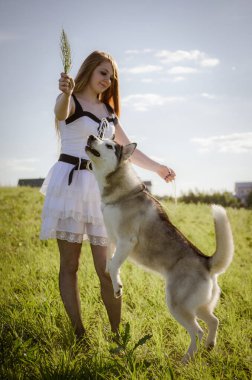 Uzun kızıl saçlı genç bir kadın çimenli bir tarlada duruyor, dalları şakacı bir şekilde kabuğunun üzerinde tutuyor. Parlak güneş ışığı yumuşak gölgeler oluşturur. Arka plandaki uzak yapı derinlik ekler.