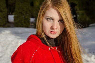 Uzun sarı saçlı, kırmızı kapüşonlu genç bir kadın, açık havada, ışıkta duruyor. Karlı arka plan kış ayarı olduğunu gösteriyor. Yüzü rahat ve kendinden emin.