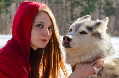 Kırmızı başlıklı genç, kızıl saçlı kadın, yoğun bir şekilde bakıyor, karlı ormanda iri yapılı tutuyor. Yumuşak güneş ışığı bulanık ağaçlara vurgu yaparak sakin doğa manzarası yaratıyor.