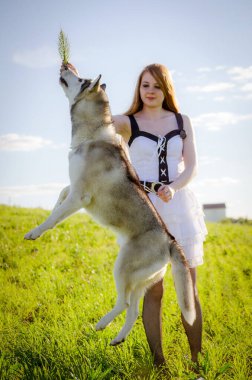 Uzun kızıl saçlı genç bir kadın çimenli bir tarlada duruyor, dalları şakacı bir şekilde kabuğunun üzerinde tutuyor. Parlak güneş ışığı yumuşak gölgeler oluşturur. Arka plandaki uzak yapı derinlik ekler.