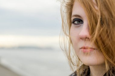 Kısmen yüzünü kapatan kızıl saçlı genç bir kadın. Ciddi ifade, soluk ten, açık renk makyaj. Bulanık gökyüzü ve bulanık deniz kenarı arkaplanı sakin bir atmosfer yaratır