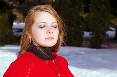 Kızıl saçlı ve soluk tenli genç bir kadın karlı ormanda duruyor. Parlak kırmızı bir üst, gözleri kapalı, ifadesini sakin. Güneş ışığı canlı renkleri ve doğal ayarı vurgular