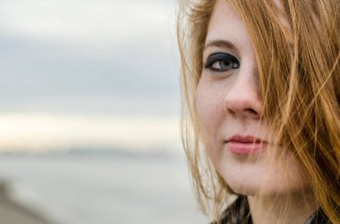 Kısmen yüzünü kapatan kızıl saçlı genç bir kadın. Ciddi ifade, soluk ten, açık renk makyaj. Bulanık gökyüzü ve bulanık deniz kenarı arkaplanı sakin bir atmosfer yaratır