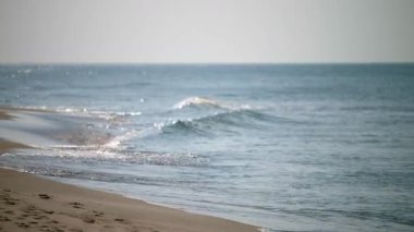 Yumuşak dalgalar açık mavi gökyüzünün altında kumlu sahile çarpıyor. Güneş ışığı su yüzeyinde parıldar, sakin ve huzurlu bir atmosfer yaratır. Kıyı şeridi boyunca görünen ayak izleri
