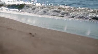 Küçük dalgaların parlak güneş ışığı altında yavaşça altın kumlu sahile ulaşmasını yakından izliyoruz. Su yansıyan ışıkla parlıyor, huzurlu ve huzurlu bir atmosfer yaratıyor.