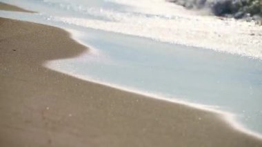 Sahil şeridine yakın çekim, yumuşak dalgalar ve pürüzsüz kum. Güneş ışığı su yüzeyinde parlak yansımalar yaratır. Huzurlu kıyı sahnesi huzuru çağrıştırıyor