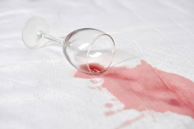 Beyaz kumaşa şarap bardağı dökülmüş, kırmızı şarap dökülmüş. Parlak ışık, şarap ve yüzey arasındaki zıtlığı vurgular. Olay yeri kaza ya da canlı bir olay olduğunu gösteriyor.