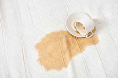Kahve fincanı beyaz kumaşın üzerine devrildi ve leke bıraktı. Sıvı düzensiz bir şekilde yayılır, koyu kahve ve açık arkaplan arasındaki doku kontrastını vurgular. Genel görünüm dökülme işaretini vurguluyor