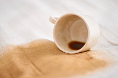 Beyaz fincanın kumaş üzerine devrilmesi kahverengi kahve lekesinin dışa doğru yayılmasıyla sonuçlandı. Işık arkaplanı koyu sıvının kontrastını arttırır. Yakın çekim açısı dağınık detayları yakalar