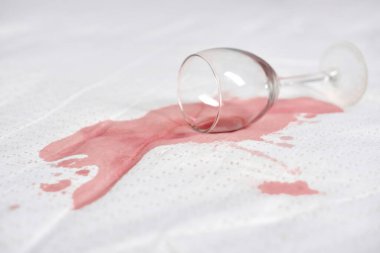 Beyaz kumaşa şarap bardağı dökülmüş, kırmızı şarap dökülmüş. Parlak ışık, şarap ve yüzey arasındaki zıtlığı vurgular. Olay yeri kaza ya da canlı bir olay olduğunu gösteriyor.