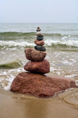 Okyanusla kumlu sahilde dengelenmiş pürüzsüz taşlar yığını. Yumuşak dalgalar yaklaşıyor, sakin bir ortam yaratıyor. Gökyüzü bulutlu ve arka plandaki kayalıklar sükuneti artırıyor