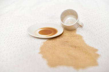 Boş bardak ve çay tabağı beyaz kumaşın üzerinde. Kahverengi sıvı yüzeye yayılır. Parlak ışıklandırma, kumaş ve kahve dökülmesi arasındaki zıtlığı artırır