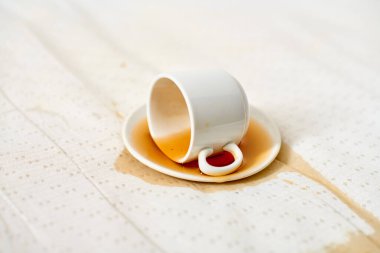 Beyaz fincan, yana yatırılmış, çay tabağının üzerinde, kahve lekeli kumaş. Yumuşak ışıklandırma, kumaş yüzeyine dökülme desenini vurgular, ince bir kontrast yaratır