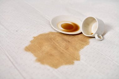 Beyaz masa örtüsünün üzerine konmuş boş kahve fincanı kahverengi sıvı leke yapıyor. Yumuşak ışıklandırma, beyaz kumaş ve koyu dökülme, kaza hissi ve sakarlık arasındaki zıtlığı vurgular.