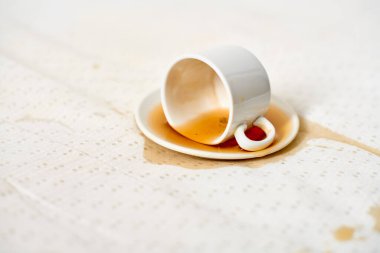 Beyaz fincan, yana yatırılmış, çay tabağının üzerinde, kahve lekeli kumaş. Yumuşak ışıklandırma, kumaş yüzeyine dökülme desenini vurgular, ince bir kontrast yaratır