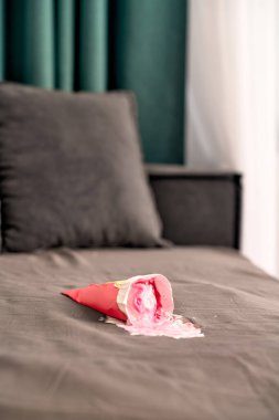 Kırmızı dondurma külahı gri koltukta uzanıyor, eriyip pembe leke yapıyor. Koyu yeşil perdeler çerçeve arka plan, kontrastı ekler. Yumuşak ışıklandırma dokuları ve renkleri geliştirir