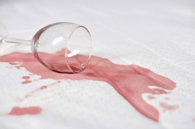 Beyaz kumaşa şarap bardağı dökülmüş, kırmızı şarap dökülmüş. Parlak ışık, şarap ve yüzey arasındaki zıtlığı vurgular. Olay yeri kaza ya da canlı bir olay olduğunu gösteriyor.