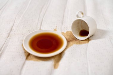 Kahve fincanı açık bej kumaşın üzerine devrilmiş. Koyu kahve çay tabağının altında havuz oluşturur. Yumuşak, dağınık ışıklandırma derindeki sıvıları vurgular