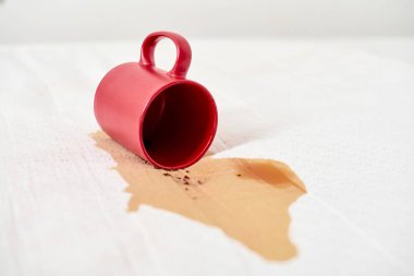 Kırmızı bardak devrildi, beyaz yüzeye kahve döküldü. Kahverengi sıvı leke yayılır ve temiz arkaplanda kontrastlar yaratır. Ekran perspektifinden çekilen sahne
