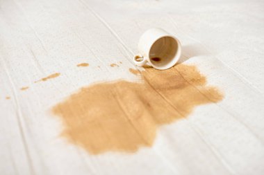 Beyaz kahve fincanı devrildi ve beyaz kumaşta kahverengi leke oluşturdu. Parlak ışıklandırma, temiz kumaş ve kahve dökülmesi arasında güçlü bir kontrastı vurguluyor. Olay yeri kaza ya da dikkatsizlik olduğunu gösteriyor.