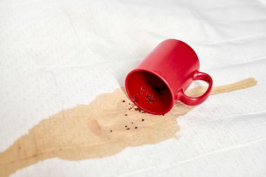 Kırmızı kupa beyaz desenli kumaşa takla atarak yayılmış bir leke yarattı. Parlak ışıklandırma, canlı kupa ve beyaz arkaplan arasındaki karşıtlığı artırarak, kaza etkisini vurguluyor