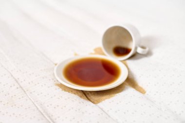 Kahve fincanı açık bej kumaşın üzerine devrilmiş. Koyu kahve çay tabağının altında havuz oluşturur. Yumuşak, dağınık ışıklandırma derindeki sıvıları vurgular