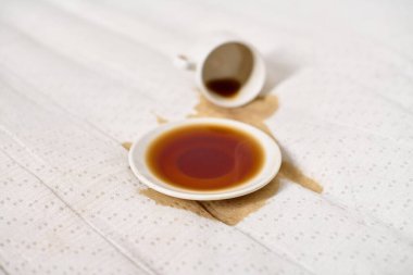 Kahve fincanı açık bej kumaşın üzerine devrilmiş. Koyu kahve çay tabağının altında havuz oluşturur. Yumuşak, dağınık ışıklandırma derindeki sıvıları vurgular