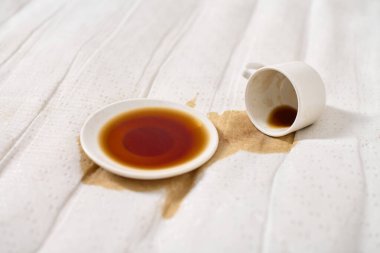 Kahve fincanı açık bej kumaşın üzerine devrilmiş. Koyu kahve çay tabağının altında havuz oluşturur. Yumuşak, dağınık ışıklandırma derindeki sıvıları vurgular