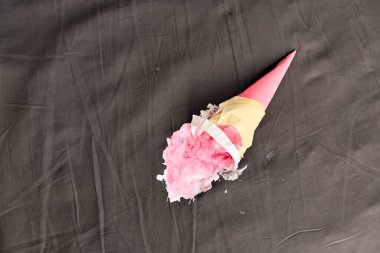 Pembe dondurma külahı karanlık yüzeyde yan yatıyordu, kısmen ezilmişti. Aydınlatma yumuşak gölgeler oluşturur, nötr gri arka planda canlı pembeyi vurgular. Beklenmedik ama neşeli anları yakalar