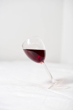 Kırmızı sıvıyla dolu cam, yumuşak beyaz arka plana eğilmiş. Sahne zarafet ve potansiyel kaos arasında, minimalist çevreler tarafından vurgulanan gerilim yaratır.