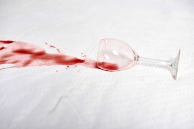 Ucundaki şarap kadehi zarif beyaz kumaşta kırmızı leke bırakır. Sıvı yayılımı katıksız kontrast gösteriyor. Görüntü yukarıdan hafif ışıklandırmayla kaydedildi