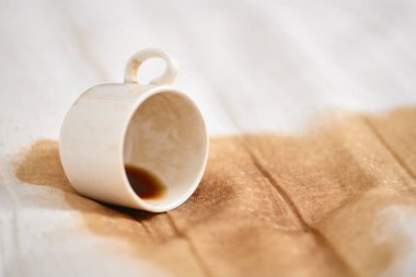 Beyaz seramik bardak yan yatmış, hafif kumaşın üzerinde küçük kahve birikintisi var. Basit kompozisyon dökülmeyi vurgular, koyu kahve ve yumuşak bej arkaplan arasında kontrastlık yaratır