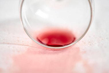 Kırmızı şarap dökülmesi beyaz kumaşta kırmızı leke yaratır. Cam yan yatmış, kazara döküldüğünü gösteriyor. Yumuşak ışıklandırma zengin şarap rengini ve hassas doku kontrastını vurgular