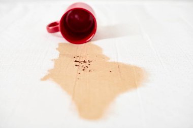 Kırmızı bardak devrilmiş, beyaz desenli yüzeye kahve dökülüyor. Kahverengi sıvı, görünür kahve telveleriyle düzensiz lekeler yaratır. Yumuşak ışık renkleri ve dokuları vurgular