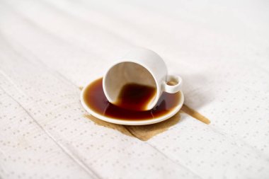Beyaz kahve fincanı beyaz noktalı kumaşın üzerine ters yatar, fincan tabağına ve kumaşa kahve döker. Parlak ışıklandırma temiz, minimalist bir atmosfer yaratır. Eğik açı, kazara döküldüğünü vurgular.
