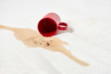 Kırmızı kupa beyaz kumaşın üzerine uzanıyor, kahve dökülüyor ve siyah leke yapıyor. Parlak ışıklandırma zıt renkleri vurgular ve karmaşanın altını çizer. Görüntü kaza hissini ve temizlik aciliyetini çağrıştırıyor