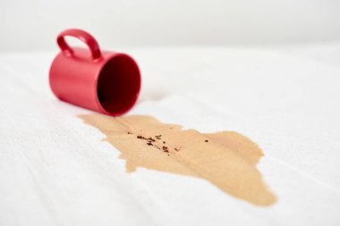 Kırmızı bardak devrildi, beyaz yüzeye kahve döküldü. Kahverengi sıvı leke yayılır ve temiz arkaplanda kontrastlar yaratır. Ekran perspektifinden çekilen sahne
