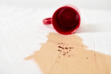 Kırmızı bardak devrildi, beyaz kumaşın üzerine kahve döküldü. Kahve taneleri yüzeye saçılmış. Parlak ışıklandırma kırmızı kupa ve beyaz arkaplan arasındaki karşıtlığı vurgular