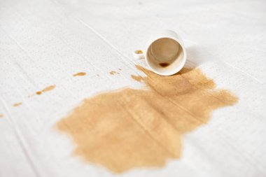 Beyaz kahve fincanı devrildi ve beyaz kumaşta kahverengi leke oluşturdu. Parlak ışıklandırma, temiz kumaş ve kahve dökülmesi arasında güçlü bir kontrastı vurguluyor. Olay yeri kaza ya da dikkatsizlik olduğunu gösteriyor.