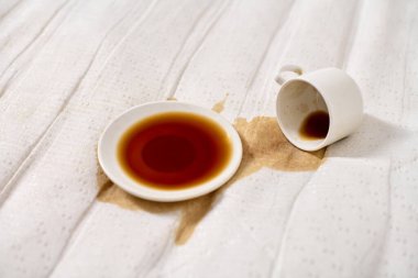 Kahve fincanı açık bej kumaşın üzerine devrilmiş. Koyu kahve çay tabağının altında havuz oluşturur. Yumuşak, dağınık ışıklandırma derindeki sıvıları vurgular