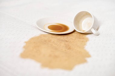 Boş bardak ve çay tabağı beyaz kumaşın üzerinde. Kahverengi sıvı yüzeye yayılır. Parlak ışıklandırma, kumaş ve kahve dökülmesi arasındaki zıtlığı artırır