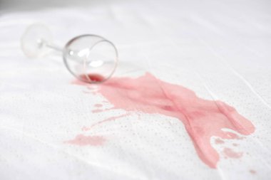 Beyaz kumaşa şarap bardağı dökülmüş, kırmızı şarap dökülmüş. Parlak ışık, şarap ve yüzey arasındaki zıtlığı vurgular. Olay yeri kaza ya da canlı bir olay olduğunu gösteriyor.