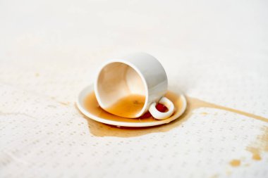 Beyaz fincan, yana yatırılmış, çay tabağının üzerinde, kahve lekeli kumaş. Yumuşak ışıklandırma, kumaş yüzeyine dökülme desenini vurgular, ince bir kontrast yaratır